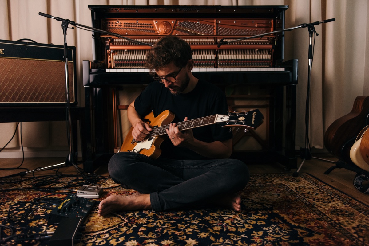 Claudio Donzelli sitting cross-legged on a rug, playing a hollow-body electric guitar